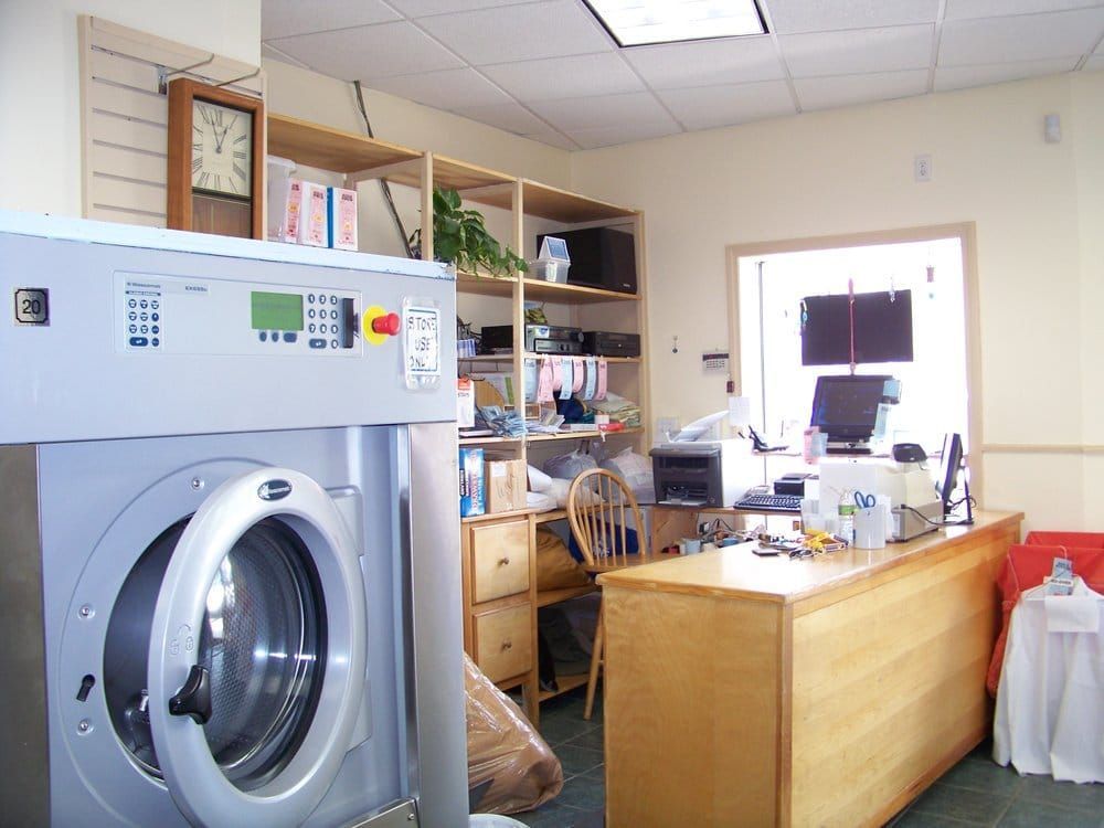 A commercial laundry with a large washer, shelves, desk, and an open doorway to an office.
