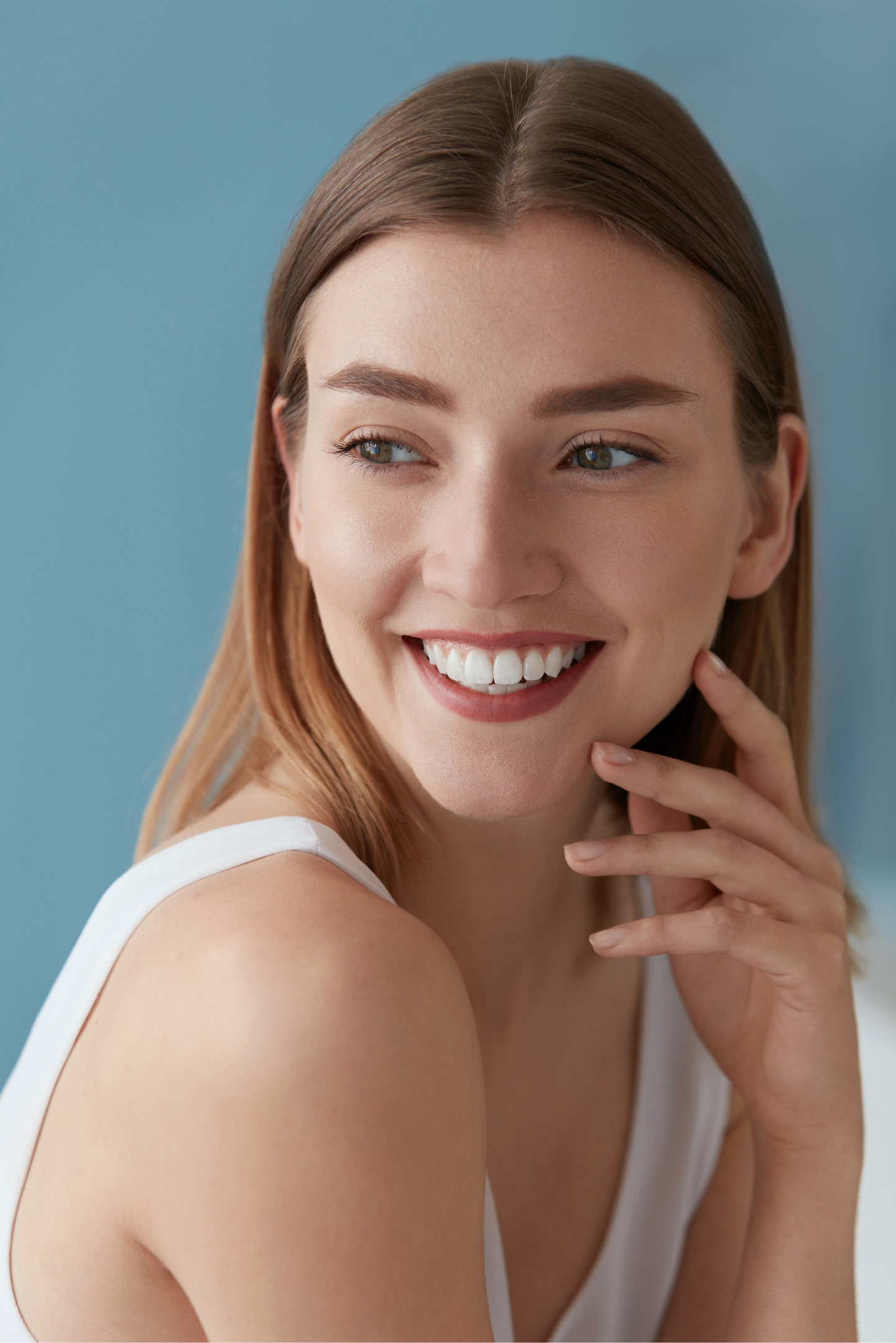 A woman in a white tank top is smiling and touching her face.