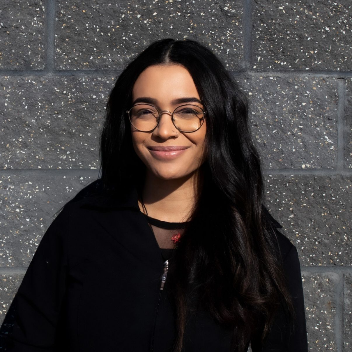 A woman in a black shirt and earrings is smiling in front of a brick wall.