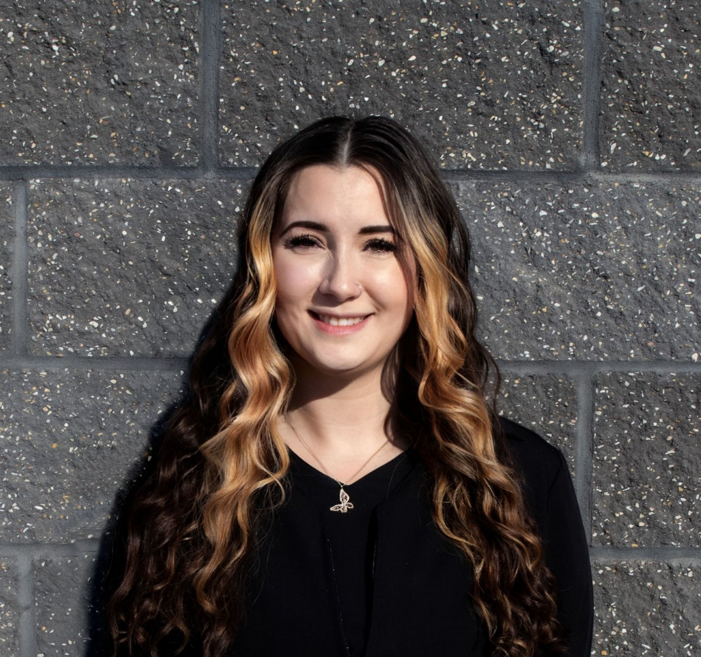 A woman in a black shirt and earrings is smiling in front of a brick wall.