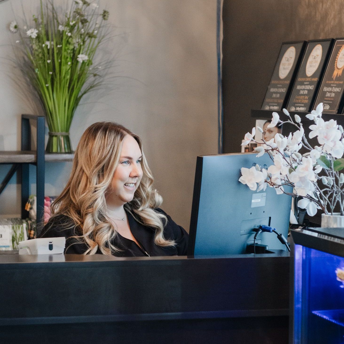 A woman is sitting at a desk in front of a computer, a receptionist at a Day Spa