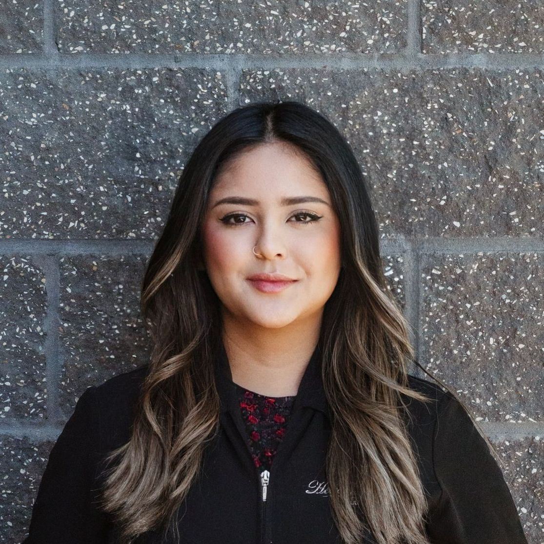 A woman in a black jacket is standing in front of a brick wall.