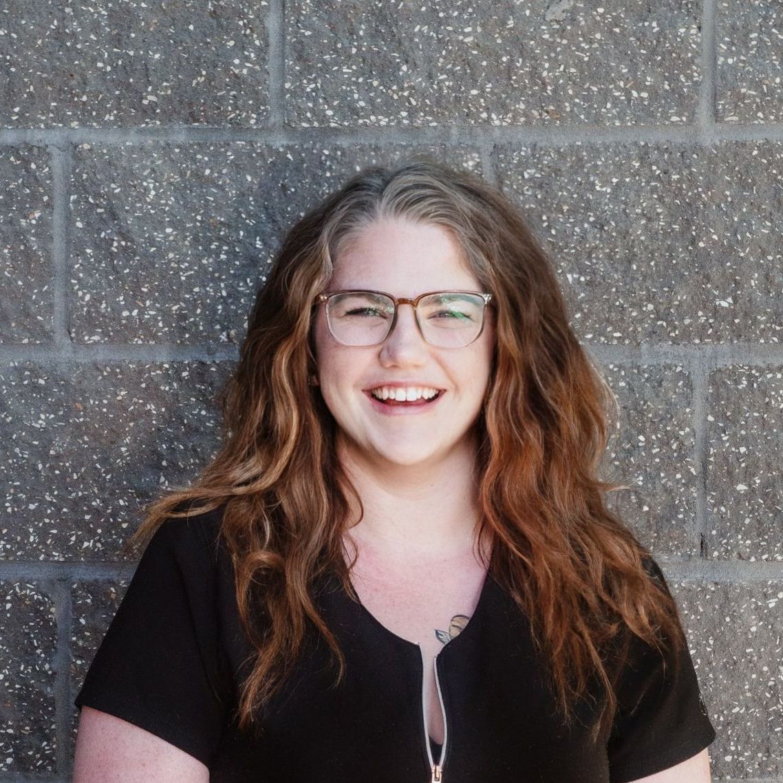 A woman wearing glasses and a black shirt is smiling in front of a brick wall
