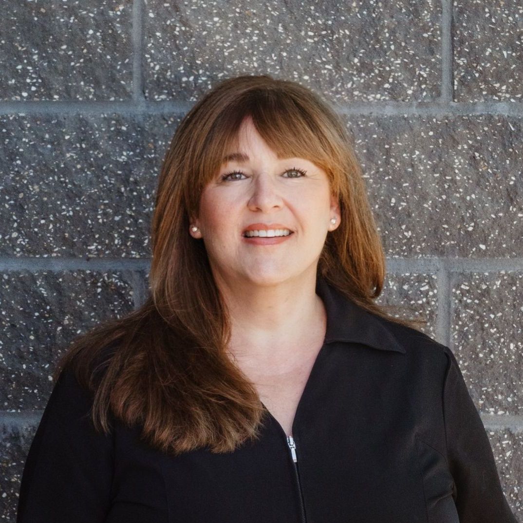 A woman in a black jacket is smiling in front of a brick wall.