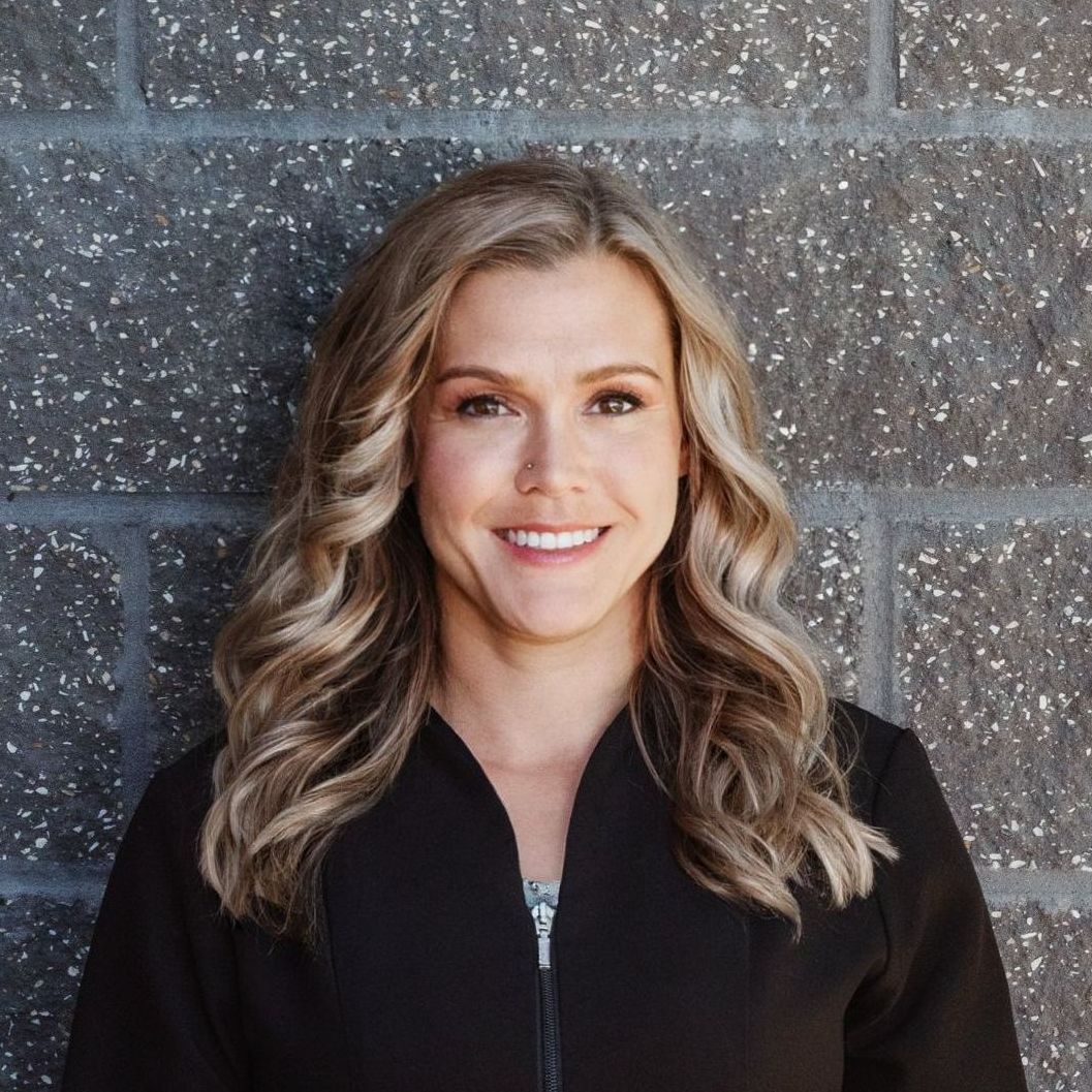A woman in a black jacket is smiling in front of a brick wall.