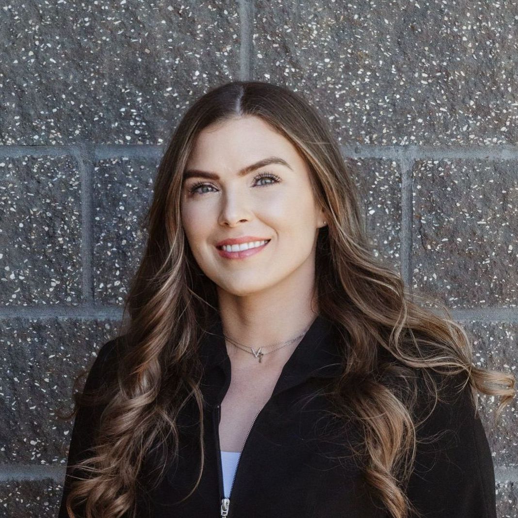 A woman in a black jacket is smiling in front of a brick wall.