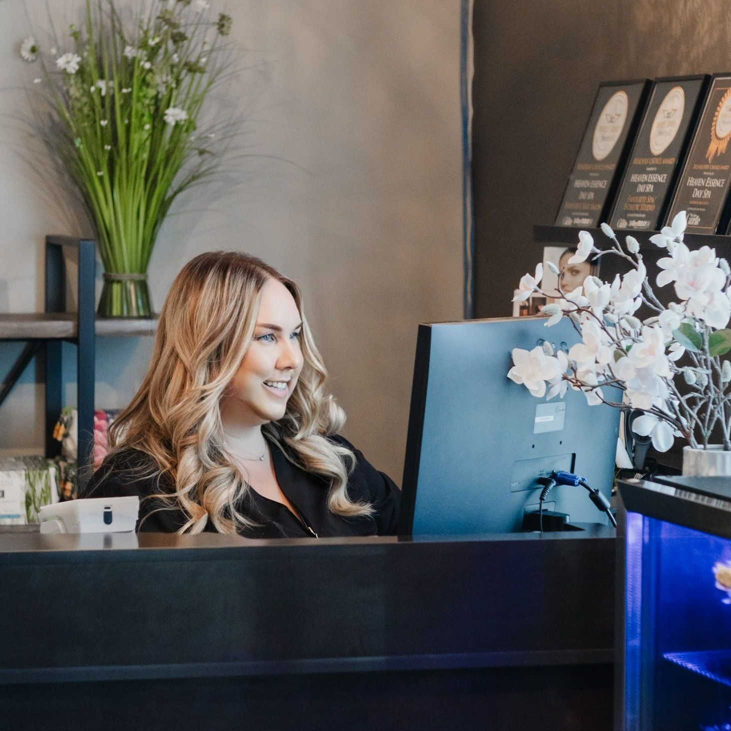 A woman is sitting at a desk in front of a computer, a receptionist at a Day Spa