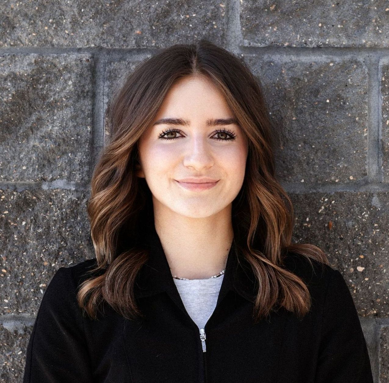 A woman in a black shirt and earrings is smiling in front of a brick wall.