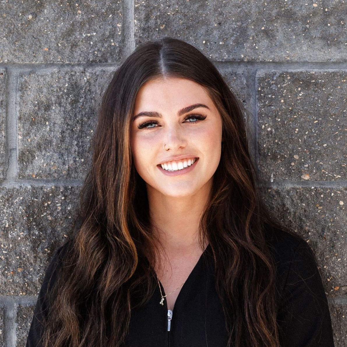 A woman in a black shirt and earrings is smiling in front of a brick wall.