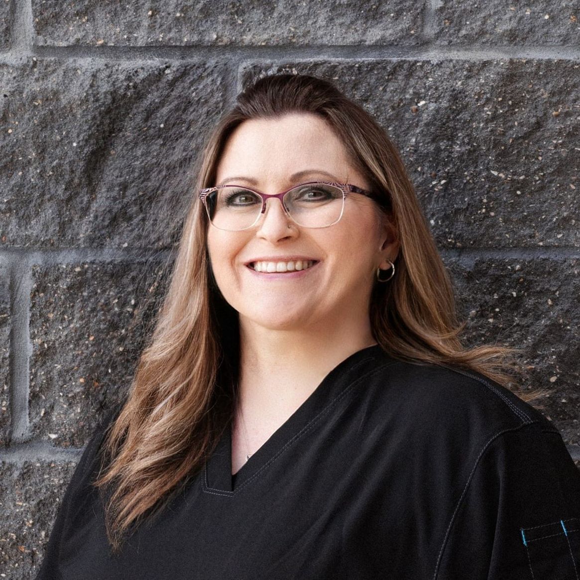 A woman in a black shirt and earrings is smiling in front of a brick wall.