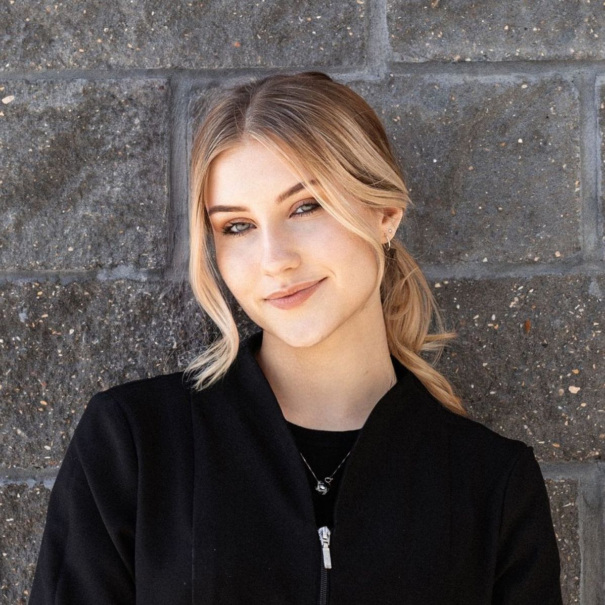 A woman in a black jacket is standing in front of a brick wall.