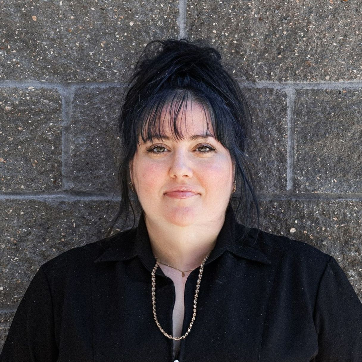 A woman in a black shirt and earrings is smiling in front of a brick wall.