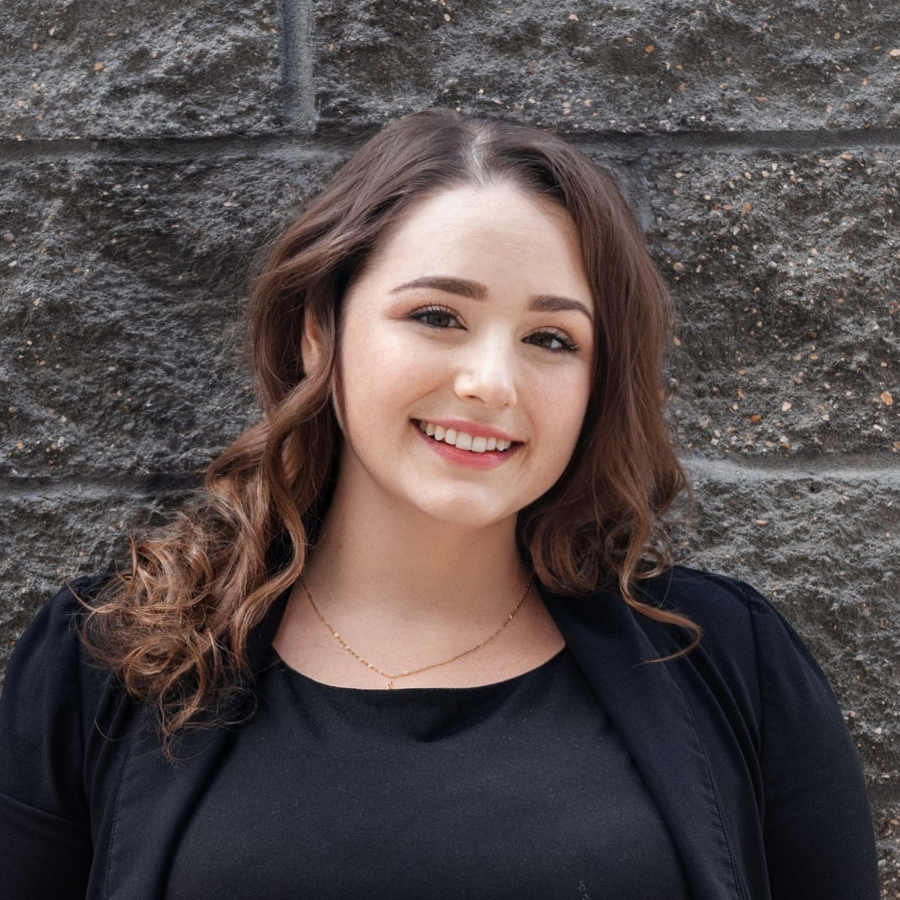 A woman in a black shirt is smiling in front of a brick wall.