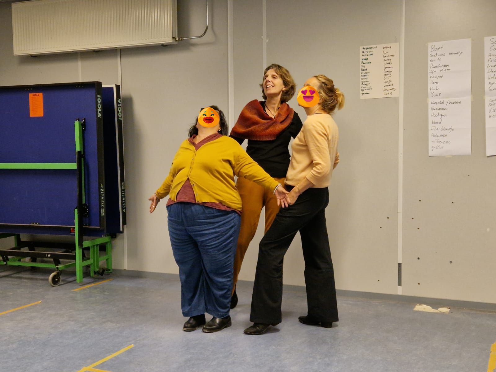 Drie mensen met oranje neuzen, hun armen uitgestrekt, kijken omhoog in een kamer met een pingpongtafel.