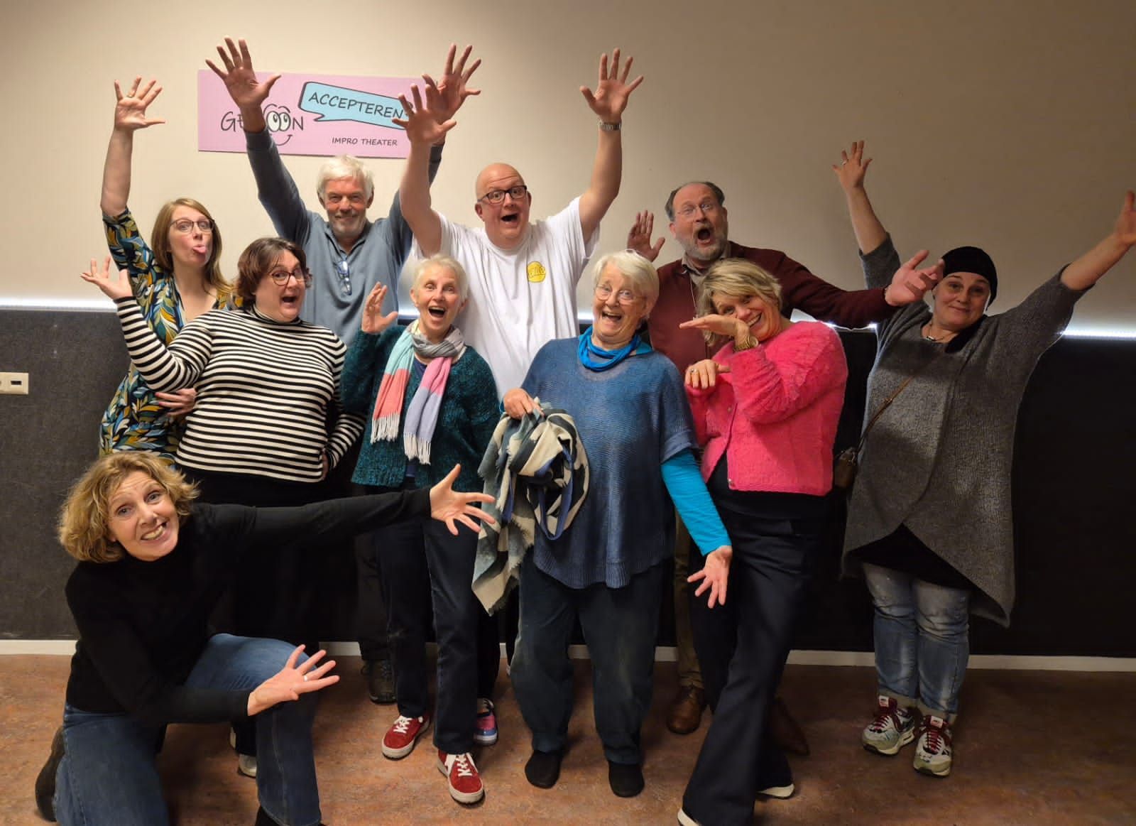 Een groep mensen poseert enthousiast binnen, met de armen omhoog en een lachend gezicht. Een roze poster is zichtbaar.