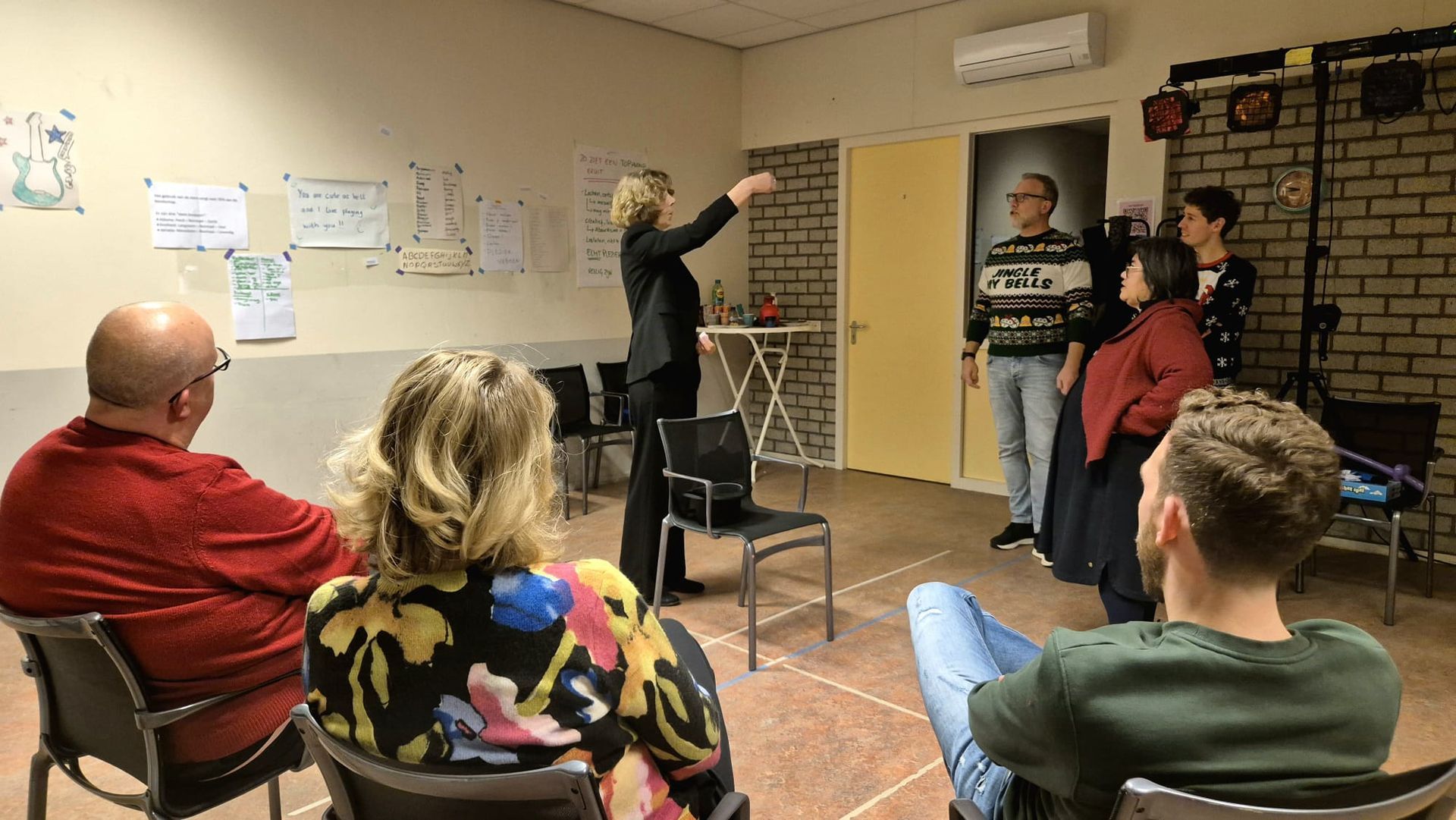 Mensen in een ruimte, onder leiding van één persoon. Sommigen zitten, anderen staan.