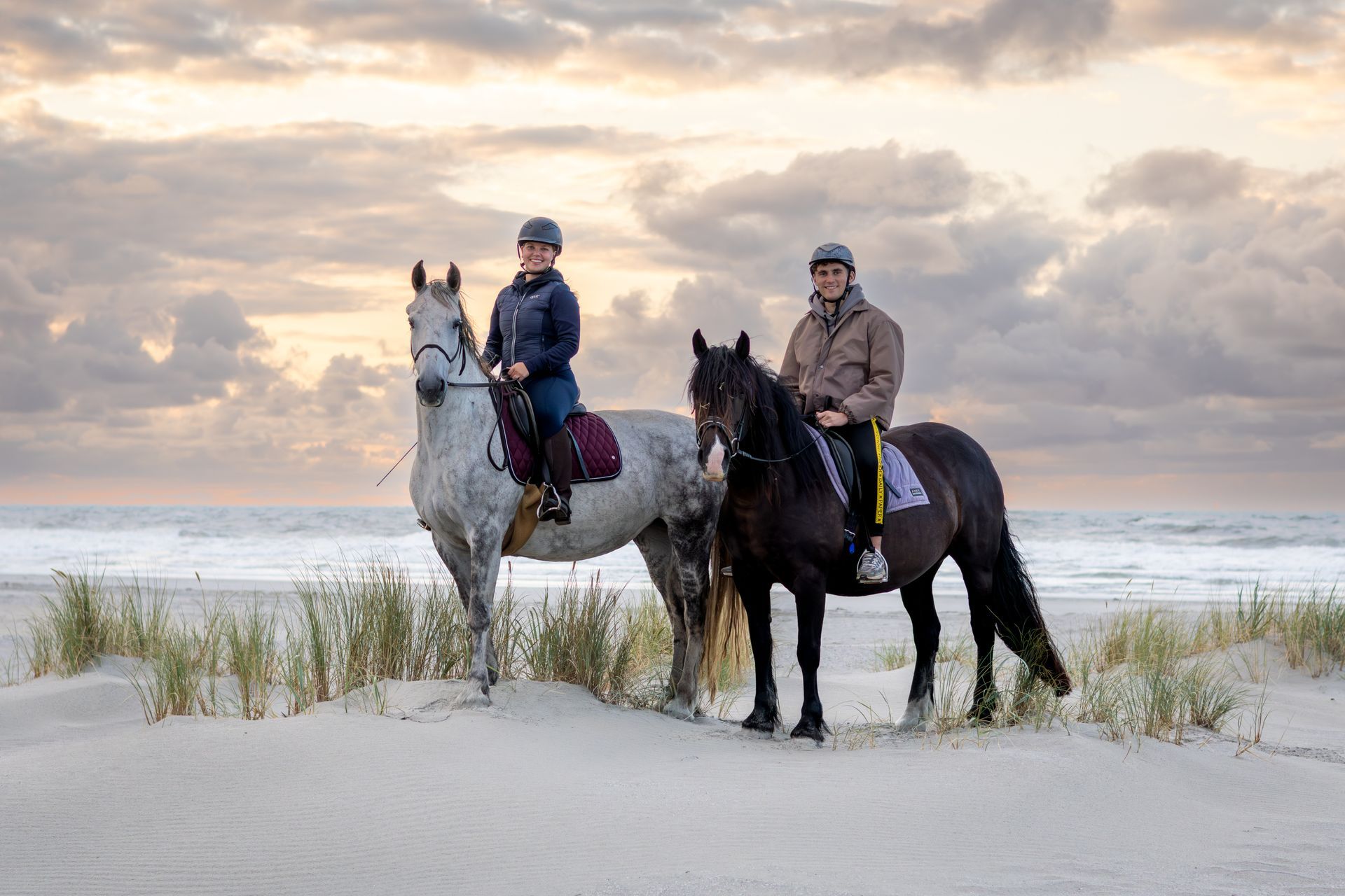 Two riders on horses at the beach, sandy dunes in foreground, ocean and cloudy sky in background.