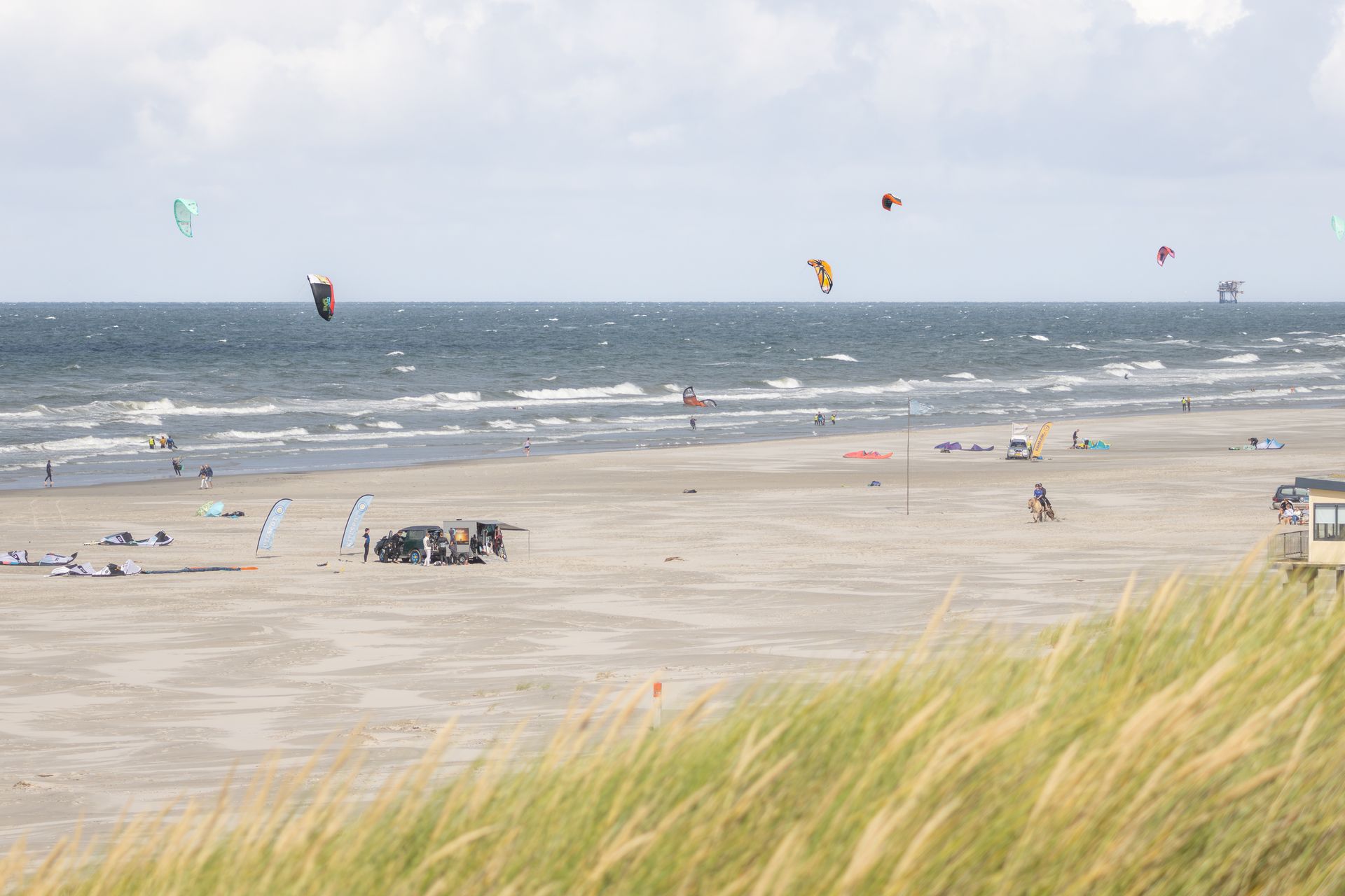 Strandtafereel met kitesurfers en mensen op het zand, de oceaan op de achtergrond, het gras op de voorgrond en de lucht is bewolkt.