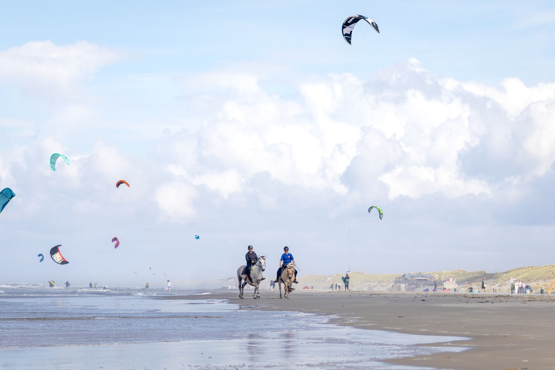 Twee mensen rijden op paarden op een zandstrand met talloze vliegers in de lucht, onder een gedeeltelijk bewolkte blauwe lucht.
