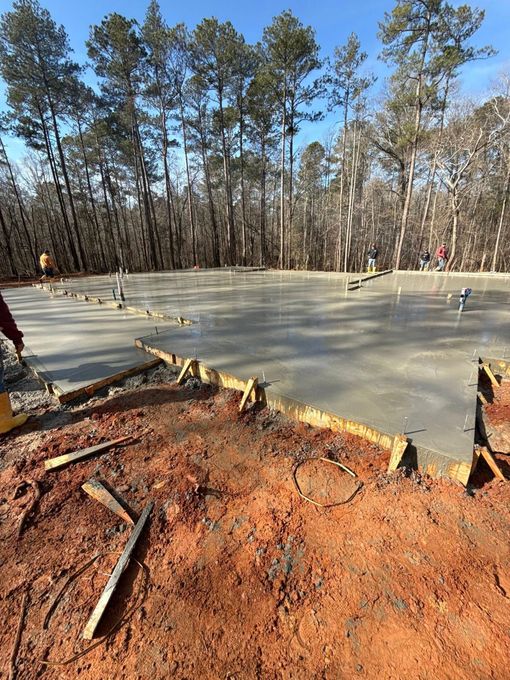 A newly poured concrete foundation surrounded by red dirt and wooden forms in a forest setting.