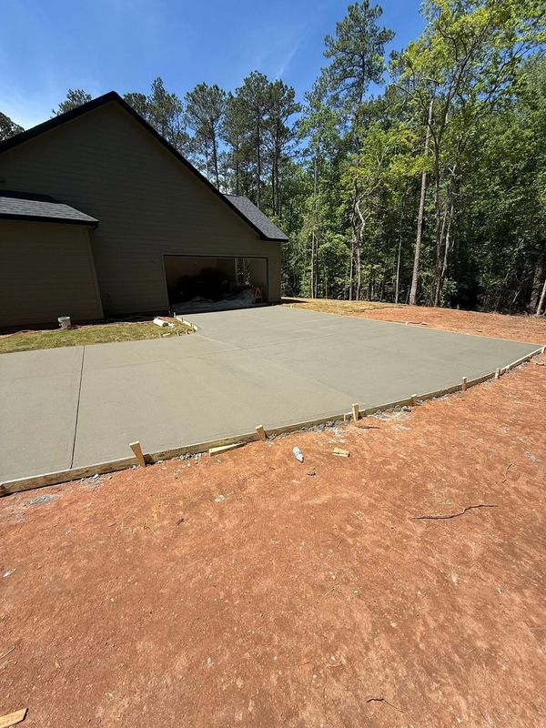 Newly poured concrete slab in a wooded area next to a house with a carport.