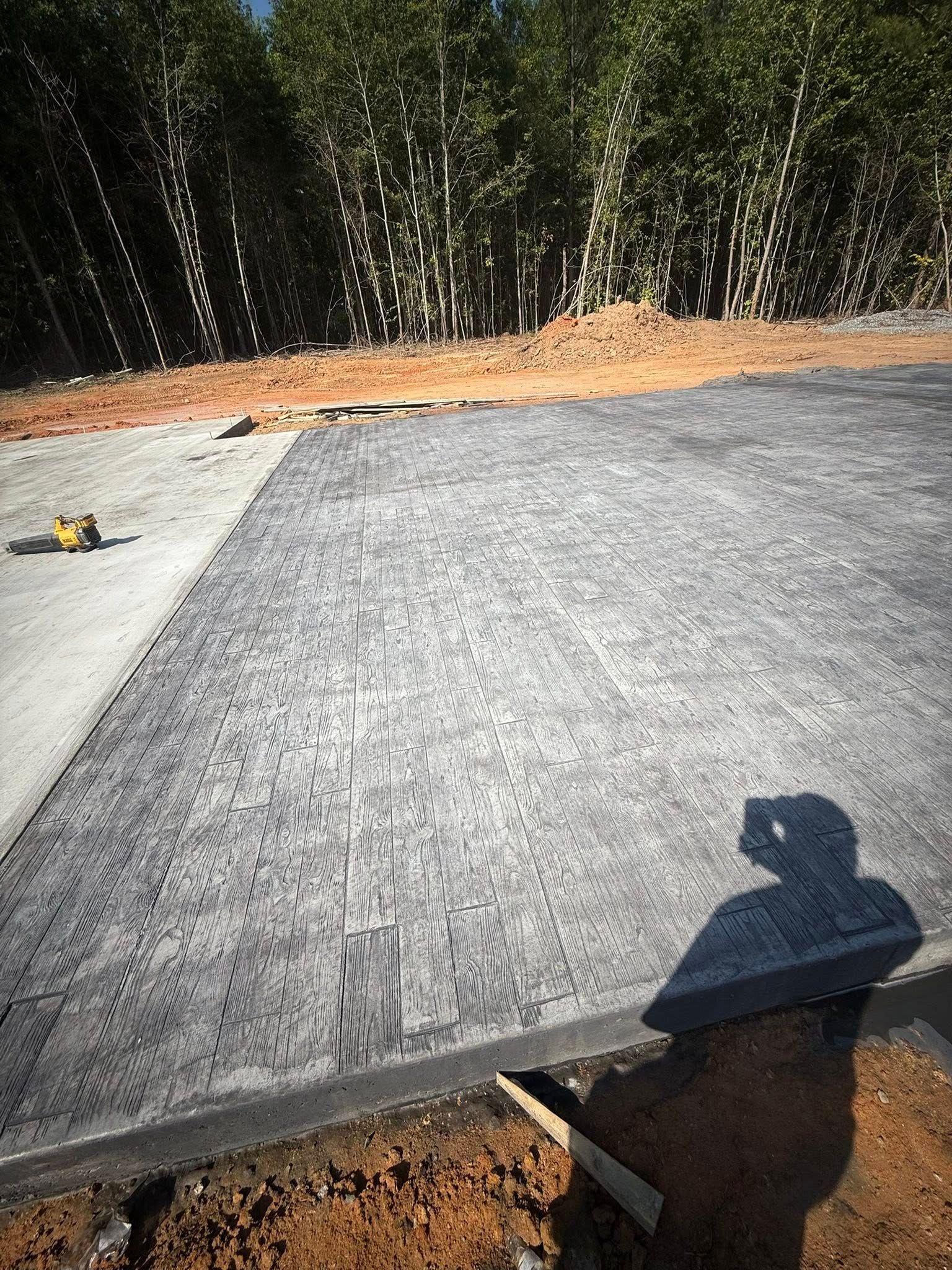 Construction site with dark paving tiles being laid. Shadow of a person holding a trowel. Trees in background.