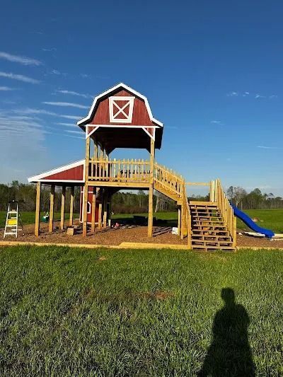 Barn-shaped wooden playground with stairs, slide, and red and white accents, set on grass with a blue sky.
