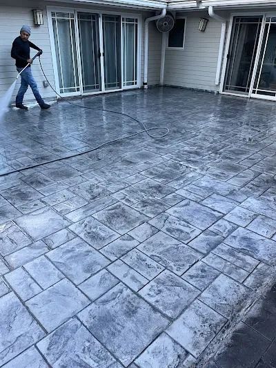 Person washing a grey stamped concrete patio with a hose in front of a house with sliding glass doors.