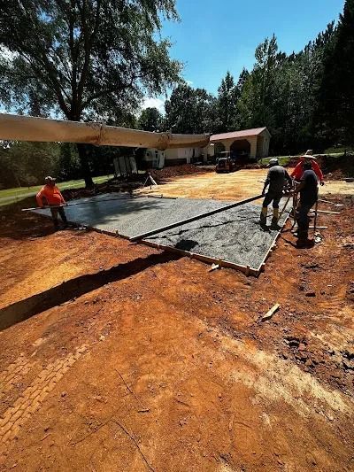 Construction workers pouring concrete foundation outdoors. Dirt, gray concrete, and trees visible.