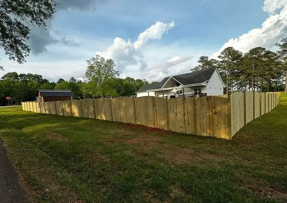 Wooden fence surrounding a house on a grassy lawn, against a backdrop of trees and a partly cloudy sky.