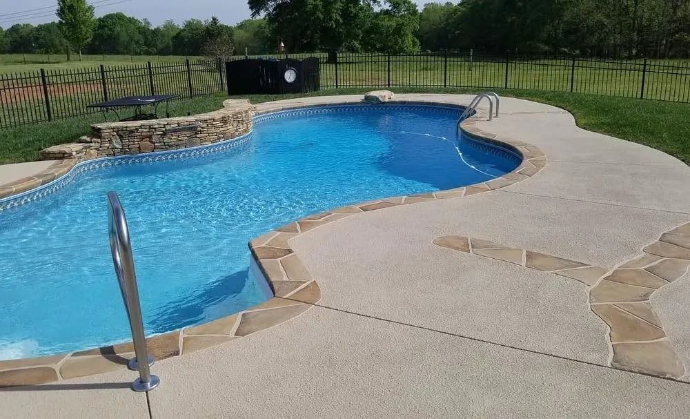 Swimming pool with blue water, surrounded by a light concrete deck with stone edging.