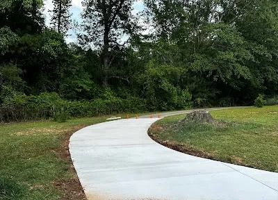 A paved path curves through a grassy area next to a wooded area.