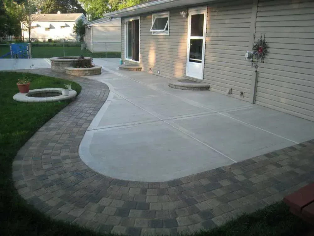 Concrete patio with brick border, leading to grass and a fire pit, next to a house.