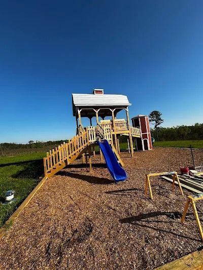 Playground structure with blue slide, wooden stairs, and red-and-white building on wood chips, under a clear blue sky.