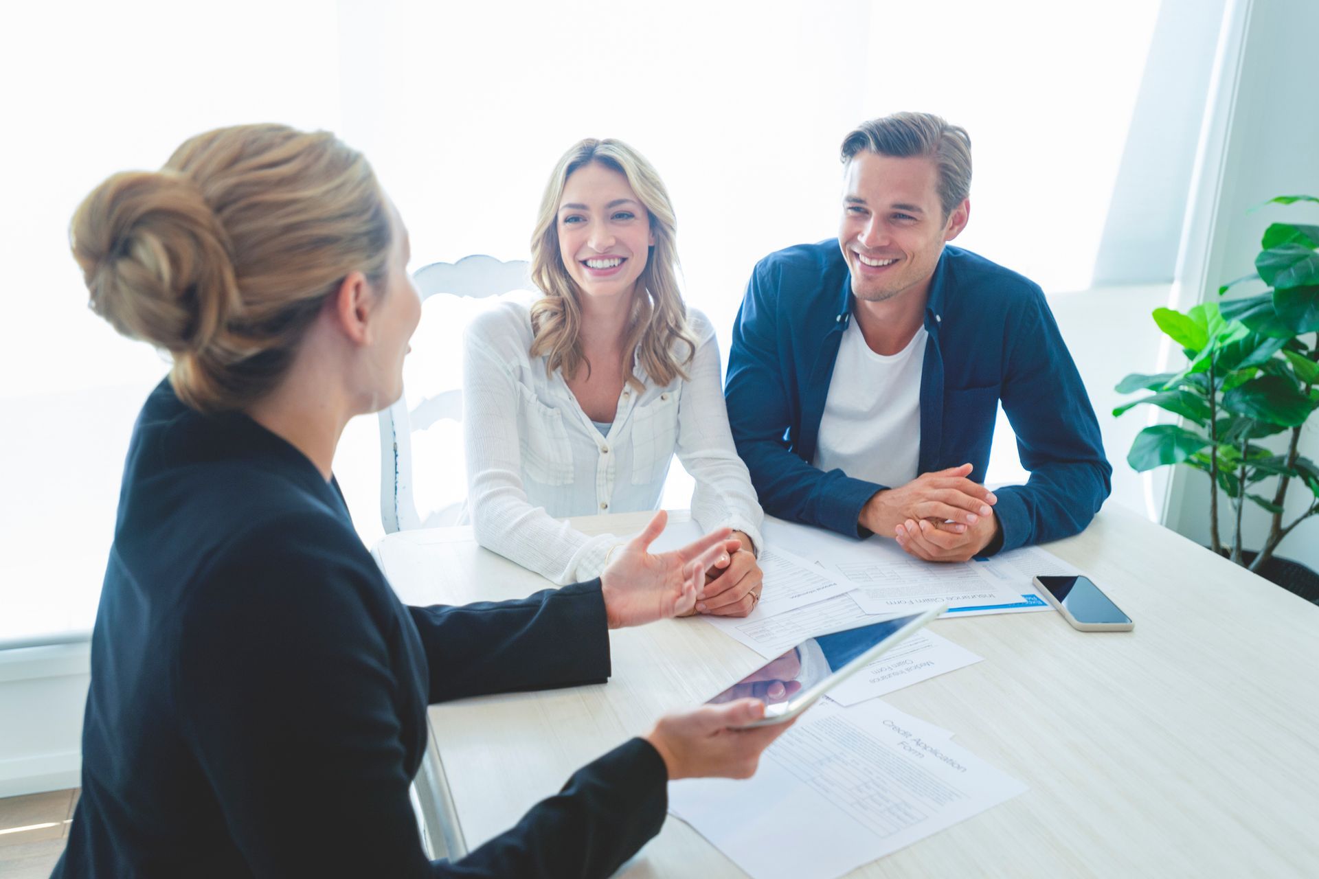 Woman in a suit talking to a smiling couple at a table; they're reviewing documents in a brightly lit room.