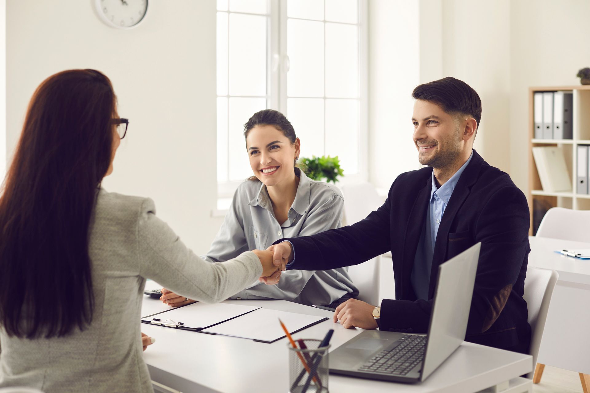 Three people at a table, one shaking hands with a man; office setting.