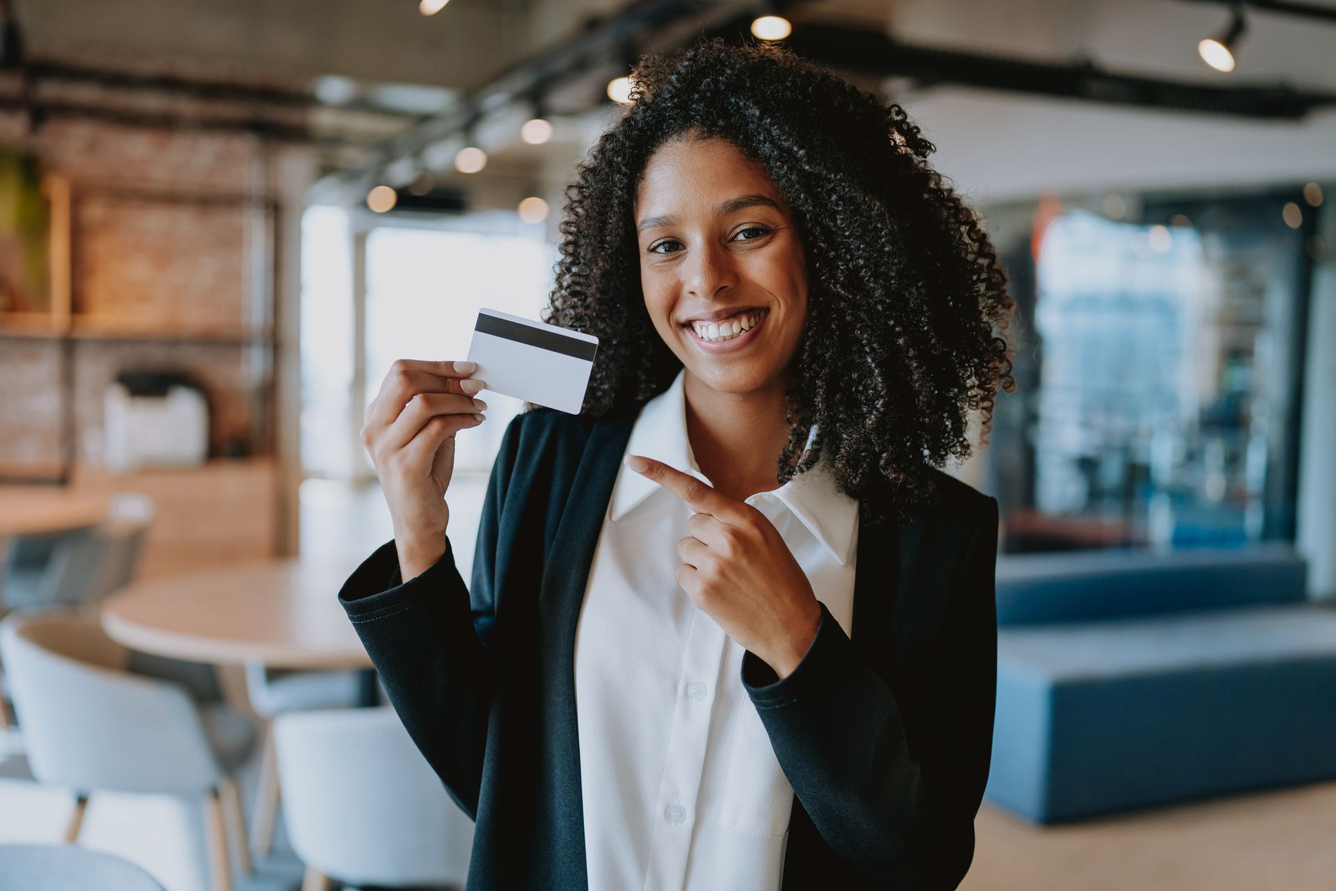 Smiling woman in business attire pointing at a credit card in a modern office setting.