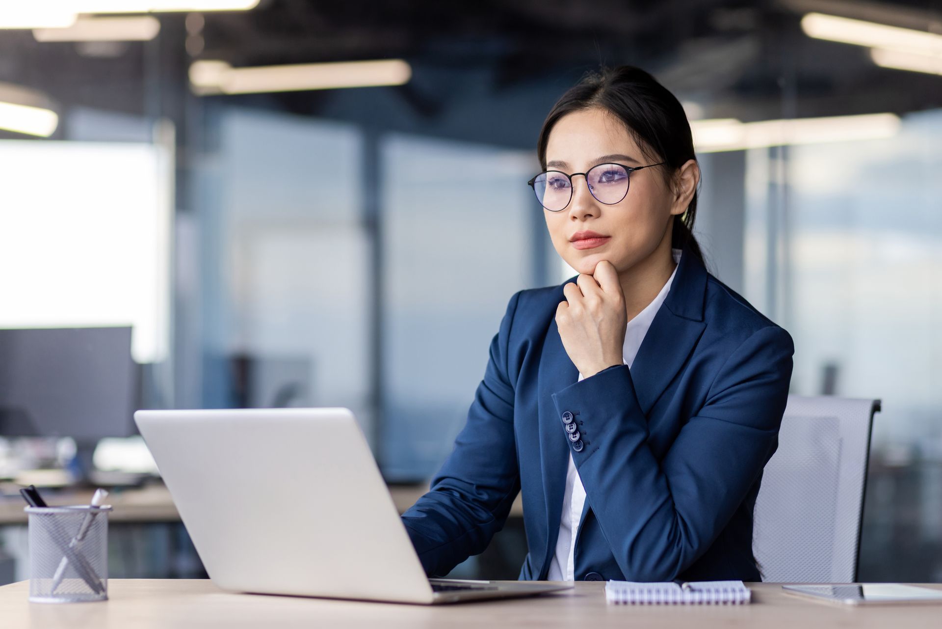 Woman in blue suit and glasses sits at desk, looking at laptop, hand on chin in office.