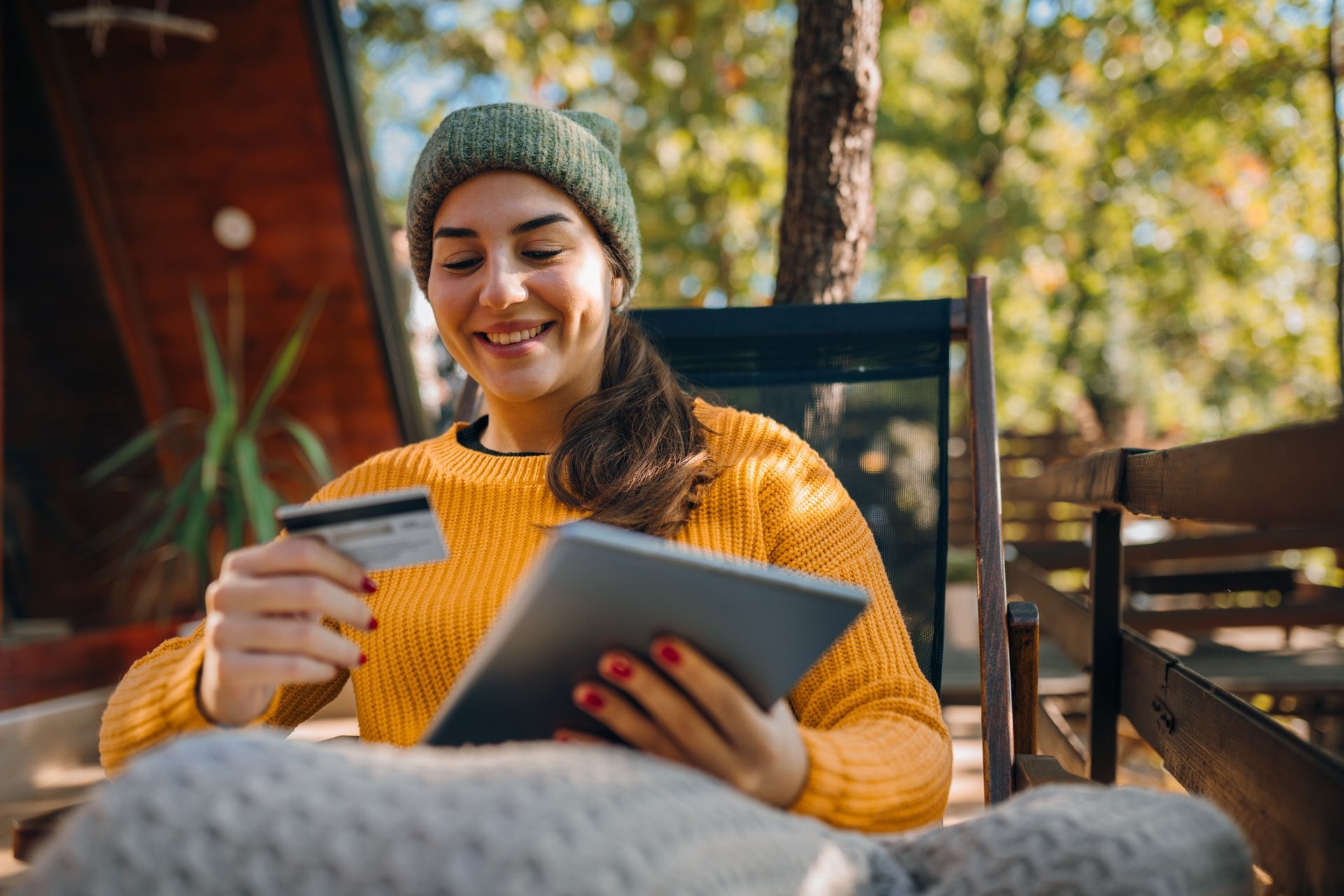 Woman in yellow sweater and beanie, shopping online outdoors with tablet and credit card.