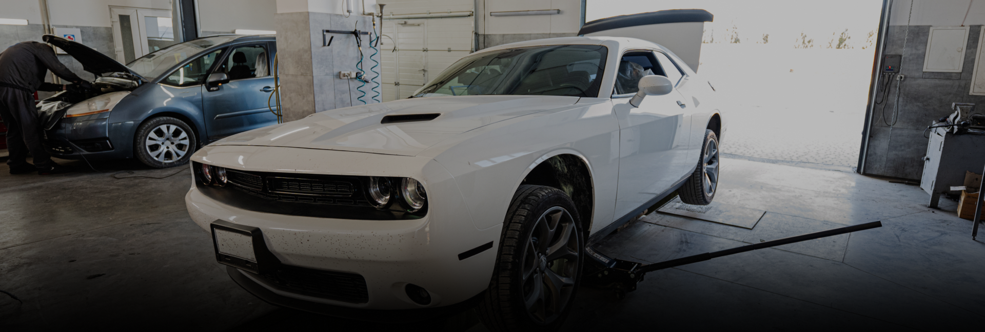 White Dodge Challenger in a garage, car being worked on by a person in the background. | Lakeside Service Center