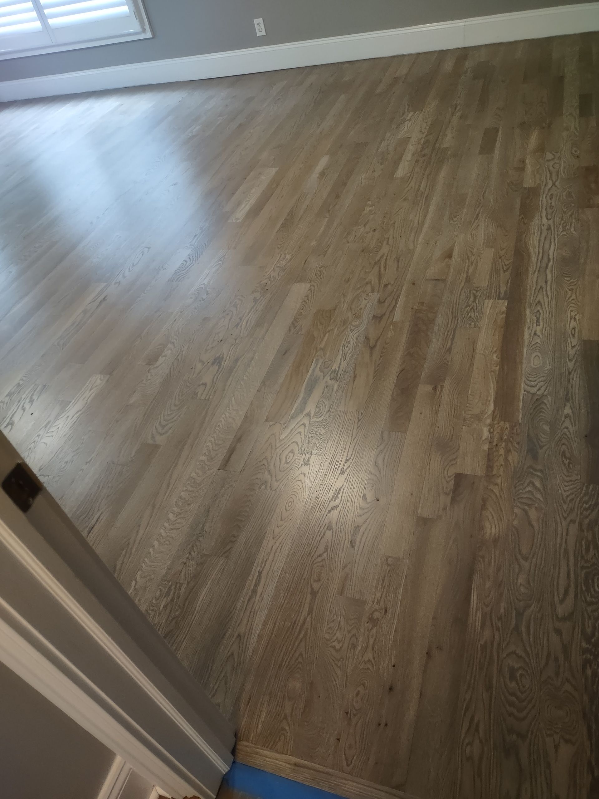 Hardwood floor with a gray-brown stain, reflecting light. The floor is in a room with a white door frame.