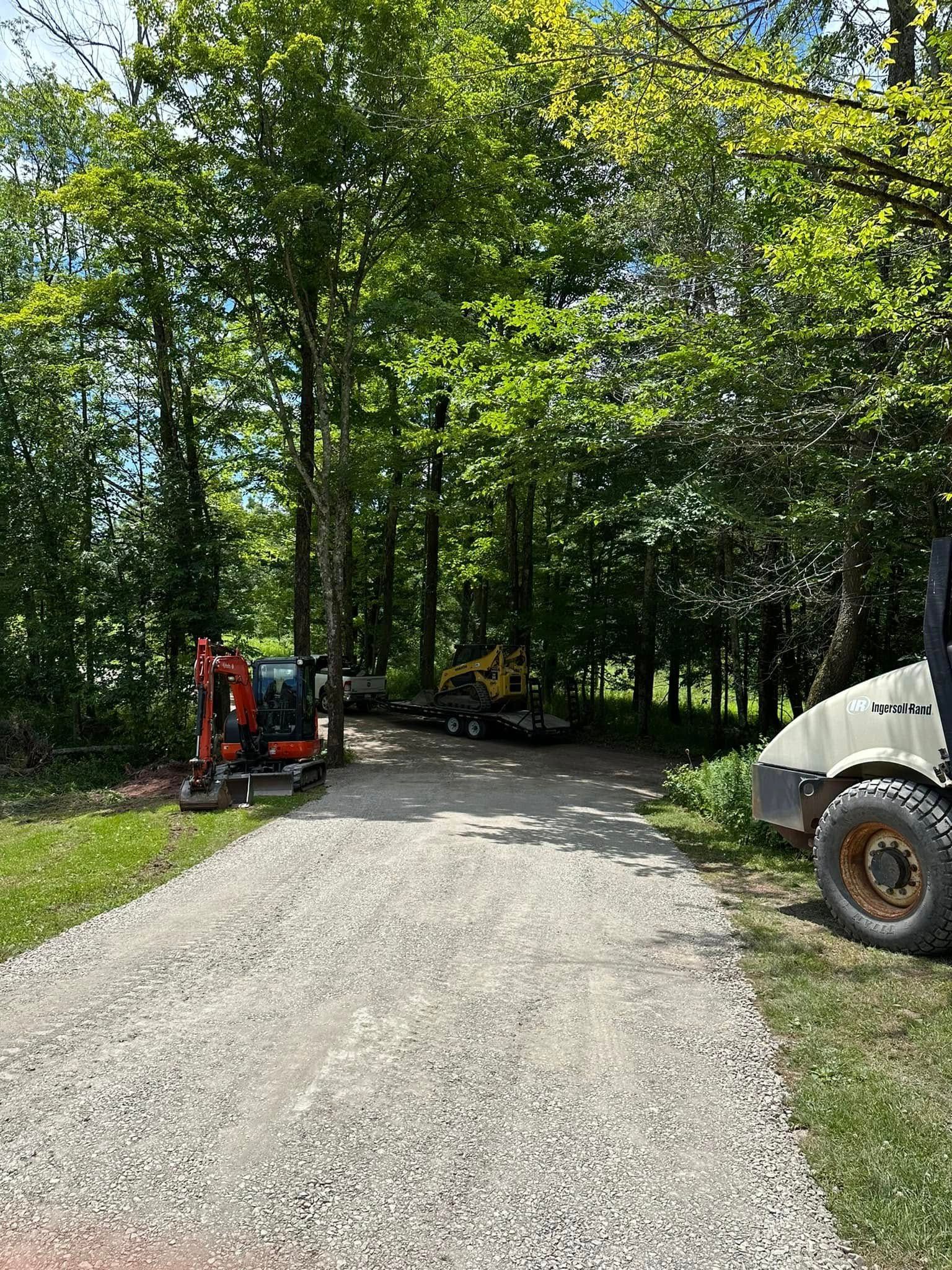 Gravel driveway leading into a wooded area with construction equipment parked on the sides.