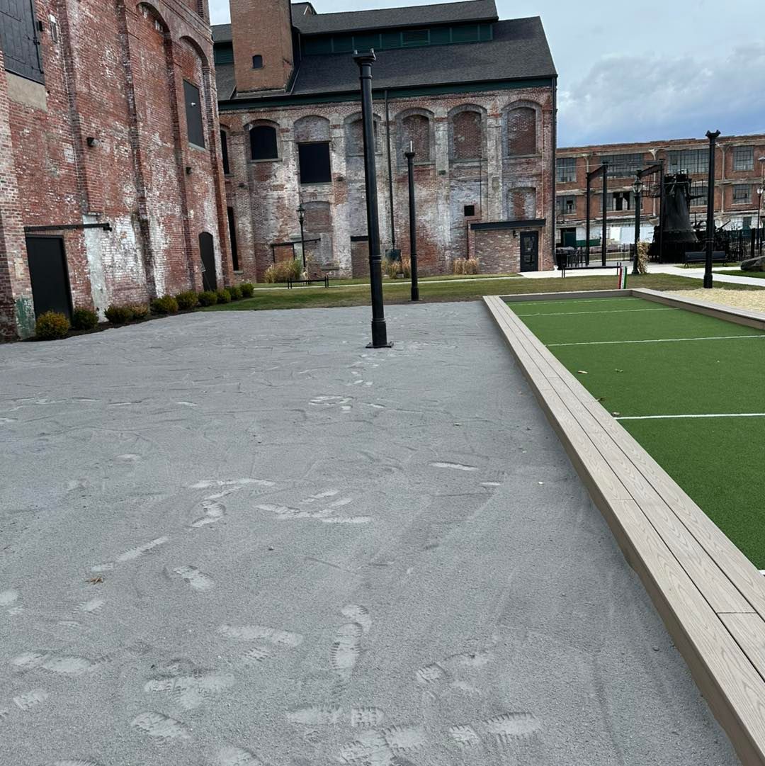 Gravel area with dark lamp posts, a synthetic lawn with a wooden border, and historic brick buildings.