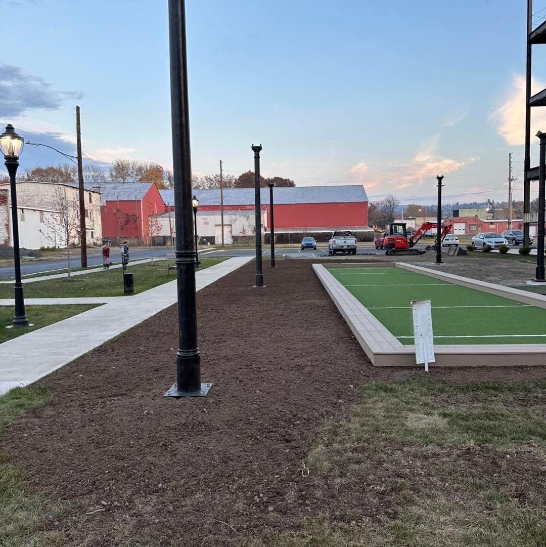 A bocce ball court in a park with artificial turf, brown mulch, and black light posts. Red barn in background.