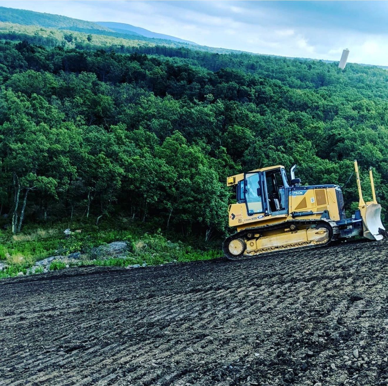 Yellow bulldozer on a freshly graded earth surface, in front of a forested hillside, under a cloudy sky.
