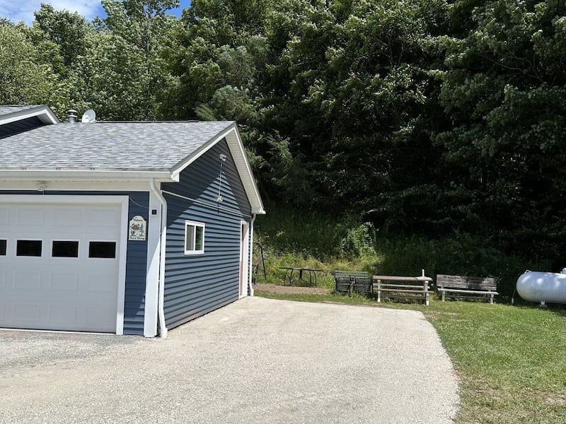 Blue building with a white garage door, gravel driveway, benches, and a large propane tank. Trees in the background.