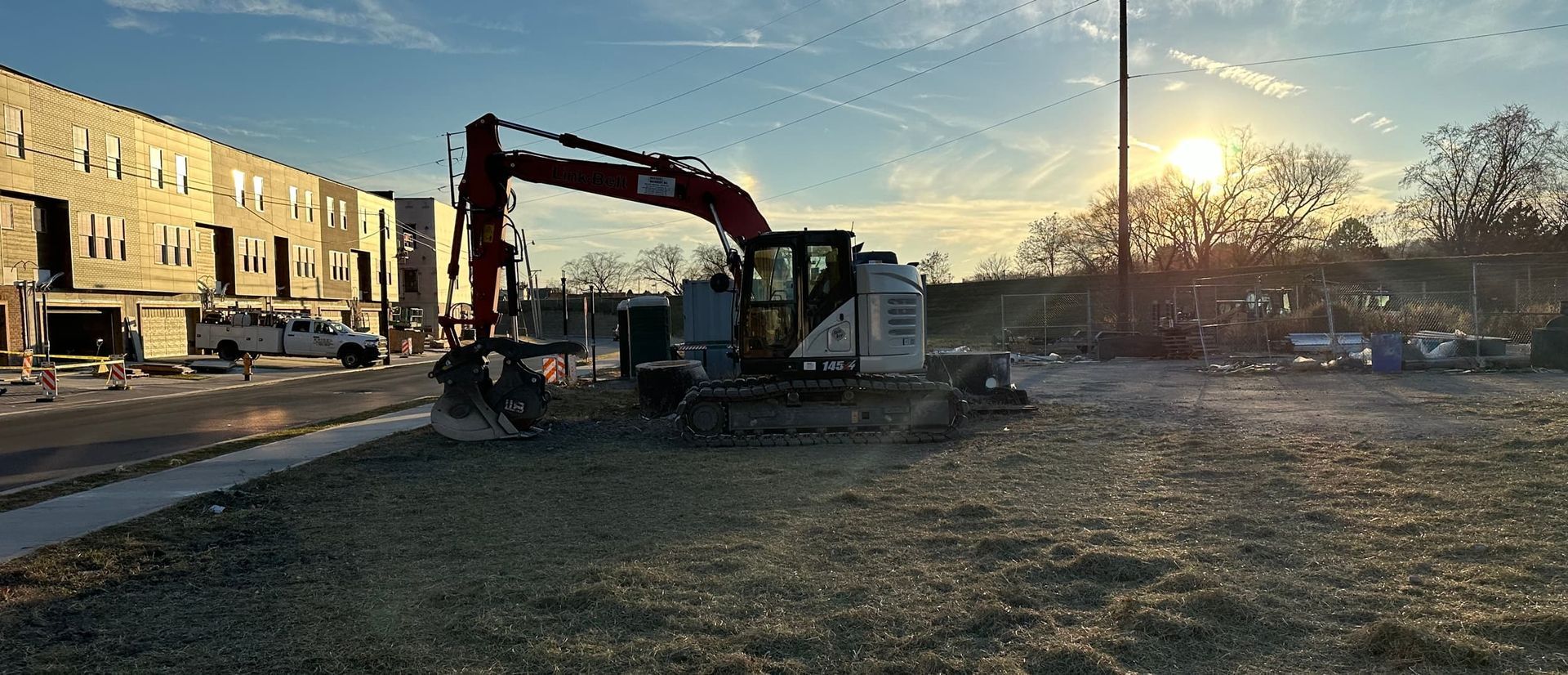 An excavator on a construction site next to a partially built building, with the sun setting.