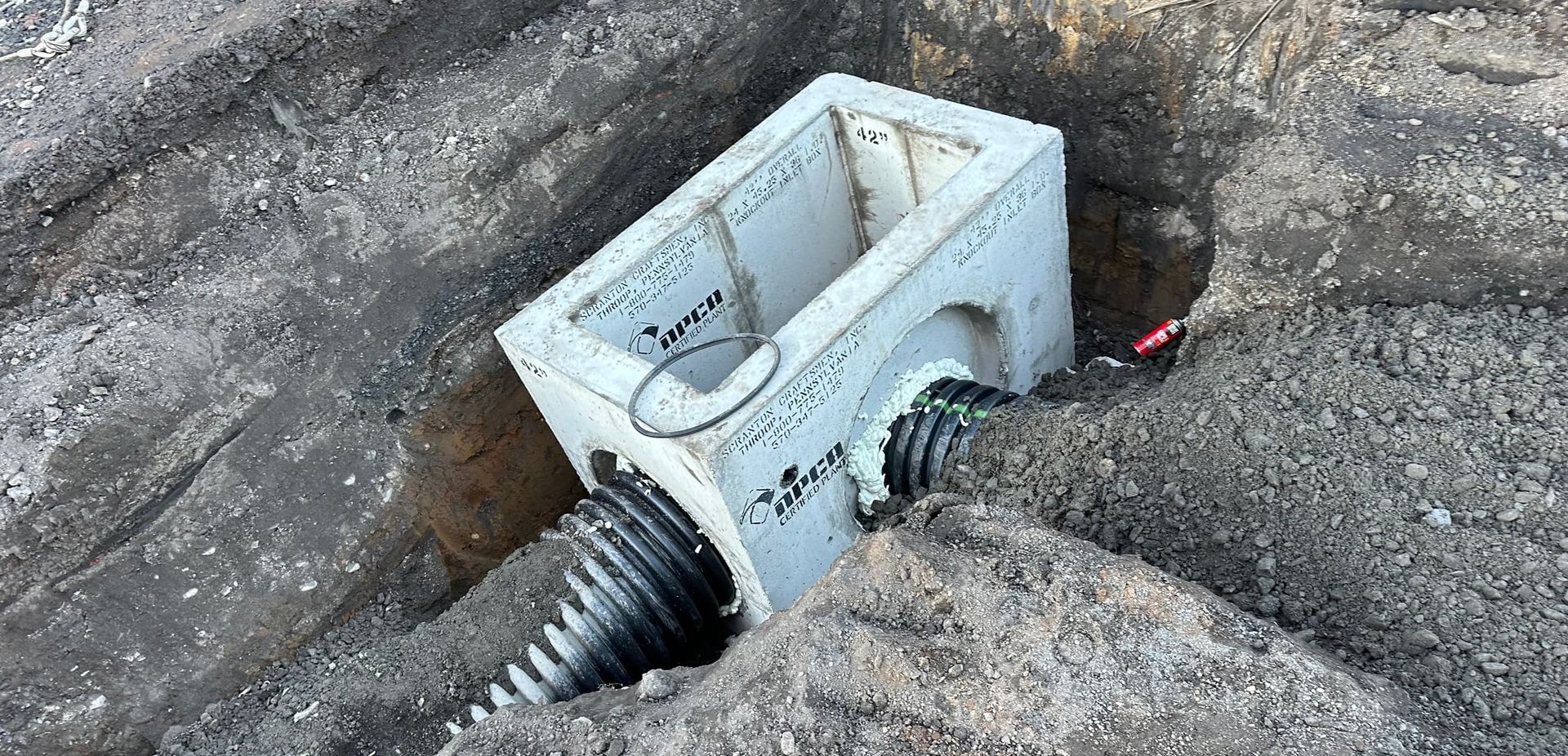 Concrete drainage box with two corrugated pipes emerging from it, set in a gravel-lined trench.
