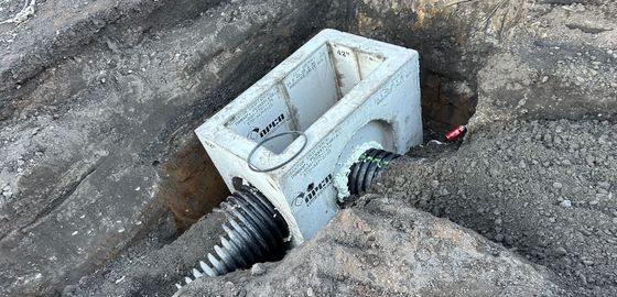 Concrete drainage box with two corrugated pipes emerging from it, set in a gravel-lined trench.