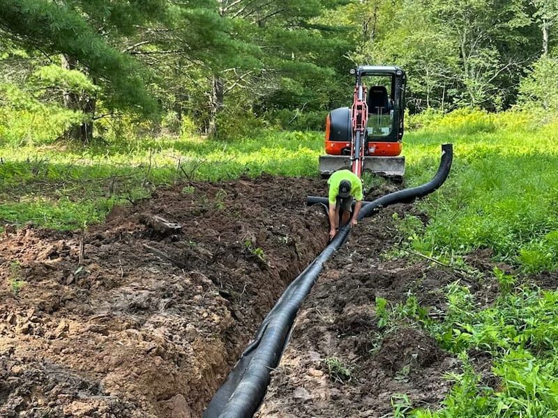 Man installing drainage pipe in a trench with an excavator in a grassy area.